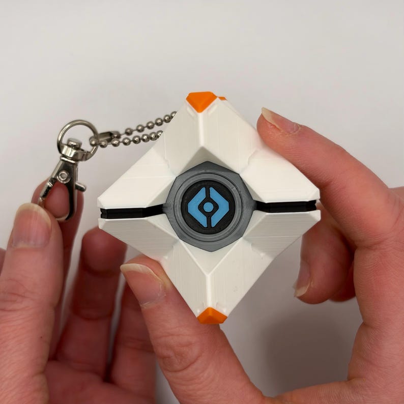 May include: A white, geometric, diamond-shaped keychain featuring orange accents at the top and bottom points. The center contains a circular gray frame surrounding a blue eye-like icon with a black center. A silver metal ball chain and clasp are attached.