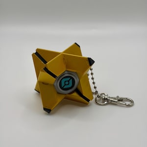 May include: A yellow, geometric, star-shaped keychain featuring a central circular element with a light blue graphic icon. The object has black accents on its pointed tips and is attached to a silver-colored metal ball chain and lobster clasp.