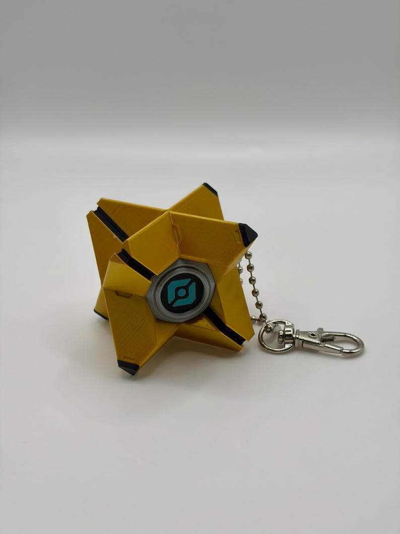 May include: A yellow, geometric, star-shaped keychain featuring a central circular element with a light blue graphic icon. The object has black accents on its pointed tips and is attached to a silver-colored metal ball chain and lobster clasp.