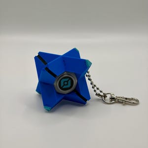 May include: A blue, geometric, star-shaped keychain featuring a central circular emblem with a black and teal graphic design. The object is attached to a silver-toned metal ball chain and a lobster clasp, showcasing a multi-faceted, angular structure.