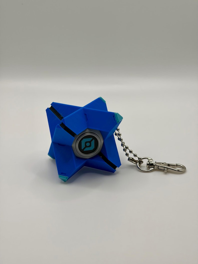 May include: A blue, geometric, star-shaped keychain featuring a central circular emblem with a black and teal graphic design. The object is attached to a silver-toned metal ball chain and a lobster clasp, showcasing a multi-faceted, angular structure.