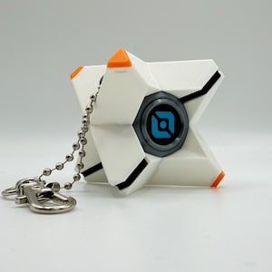 May include: A white, star-shaped keychain featuring a central circular element with a blue and black geometric logo. The object has orange accents on the tips and black linear details, attached to a silver-toned metal ball chain and lobster clasp.