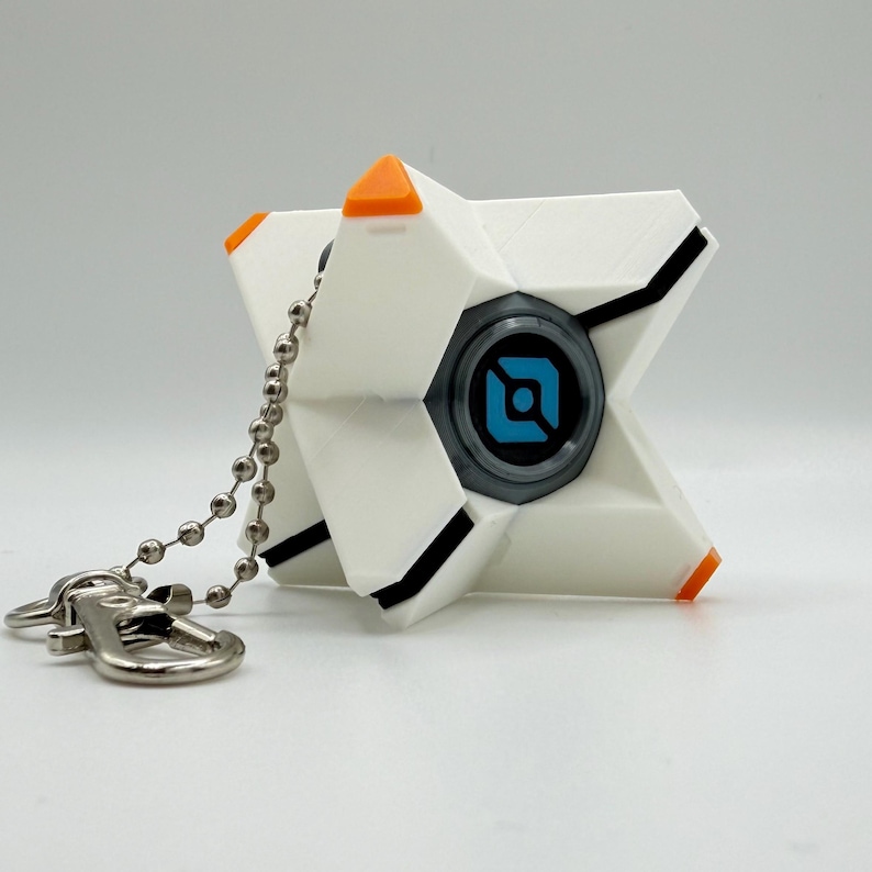 May include: A white, star-shaped keychain featuring a central circular element with a blue and black geometric logo. The object has orange accents on the tips and black linear details, attached to a silver-toned metal ball chain and lobster clasp.
