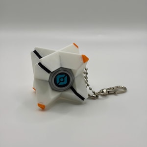 May include: A white, star-shaped geometric keychain featuring orange accents on the tips and a central circular emblem with a blue and black graphic design. The object is attached to a silver-toned metal ball chain with a lobster claw clasp.