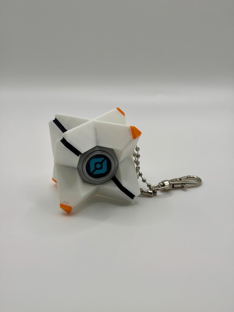 May include: A white, star-shaped geometric keychain featuring orange accents on the tips and a central circular emblem with a blue and black graphic design. The object is attached to a silver-toned metal ball chain with a lobster claw clasp.