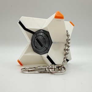 May include: A white, star-shaped geometric keychain featuring a central dark gray circular element with a horizontal slot. The points of the star are tipped with bright orange accents. A silver metal ball chain and lobster clasp are attached.
