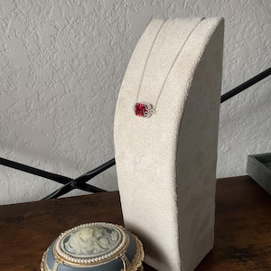May include: A silver chain necklace with a red rectangular pendant on a beige velvet display stand. A gray music box with a white cameo is in the foreground.