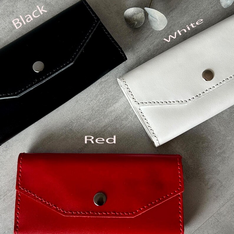 May include: Three leather wallets in black, white, and red. Each wallet has a flap closure with a silver button. The word "Black" is above the black wallet, "White" is above the white wallet, and "Red" is above the red wallet.