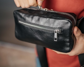 May include: A black leather fanny pack with a silver zipper and a silver zipper pull. The bag is being held by a person.