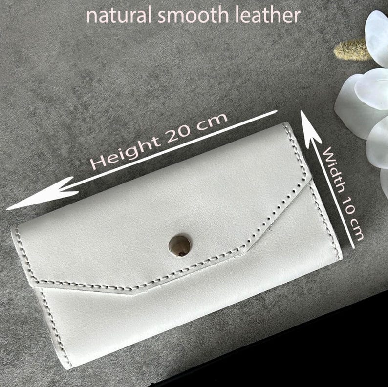 May include: A white leather wallet with a button closure. The wallet is 7.9 inches in height and 3.9 inches in width. The text "natural smooth leather" is visible at the top. The wallet has a stitched design.