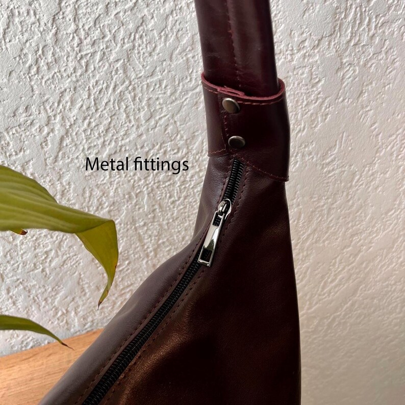 May include: A burgundy leather handbag with a silver zipper and metal fittings. The bag has a curved shape and a long strap. The text "Metal fittings" is visible on the image.