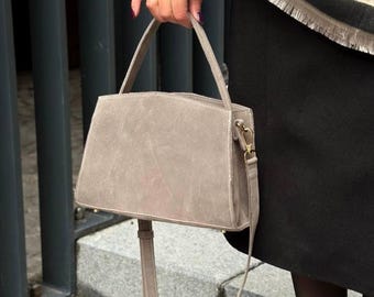 May include: A light grey suede handbag with a structured rectangular shape. The bag features a top handle and a long, adjustable shoulder strap. Gold-coloured hardware accents the bag's design, adding a touch of elegance to this everyday accessory.
