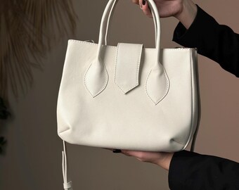 May include: A white leather tote bag with a top handle and a detachable shoulder strap. The bag has a flap closure with a buckle and decorative stitching.