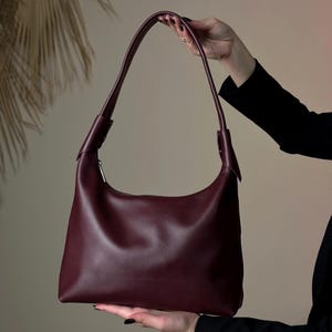 May include: A burgundy leather shoulder bag with a curved design. The bag has a single shoulder strap and a silver zipper. The bag is held up by a person wearing a black long-sleeve shirt.