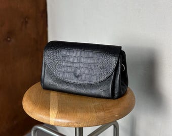 May include: A black leather crossbody bag with a crocodile-skin texture flap closure. The bag is rectangular with a rounded flap and a small strap. The bag is sitting on a wooden stool.