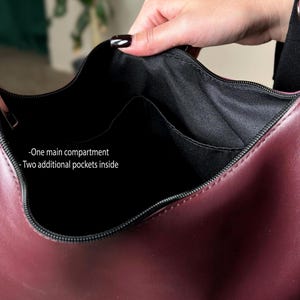 May include: Burgundy-colored handbag with a black interior. The bag features a main compartment and two additional pockets inside. The zipper is black, and the bag is held open by a hand.