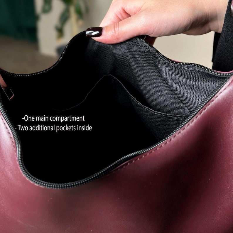 May include: Burgundy-colored handbag with a black interior. The bag features a main compartment and two additional pockets inside. The zipper is black, and the bag is held open by a hand.
