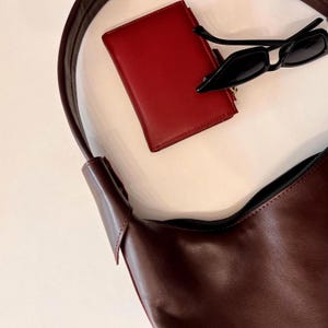 May include: A burgundy leather shoulder bag with a zipper, a red wallet, and black sunglasses. The bag has a long strap. The wallet is rectangular. The sunglasses have a rectangular frame.