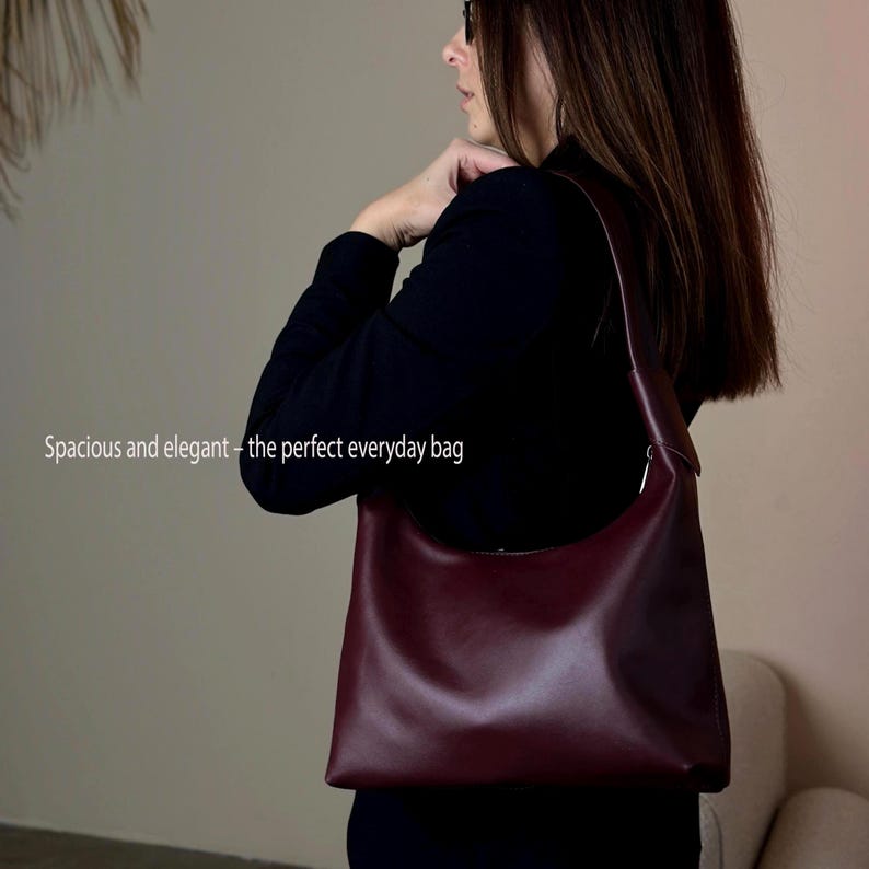 May include: A burgundy leather shoulder bag with a curved design. The bag has a long strap and a silver-toned zipper. The text "Spacious and elegant - the perfect everyday bag" is visible on the left side of the image.