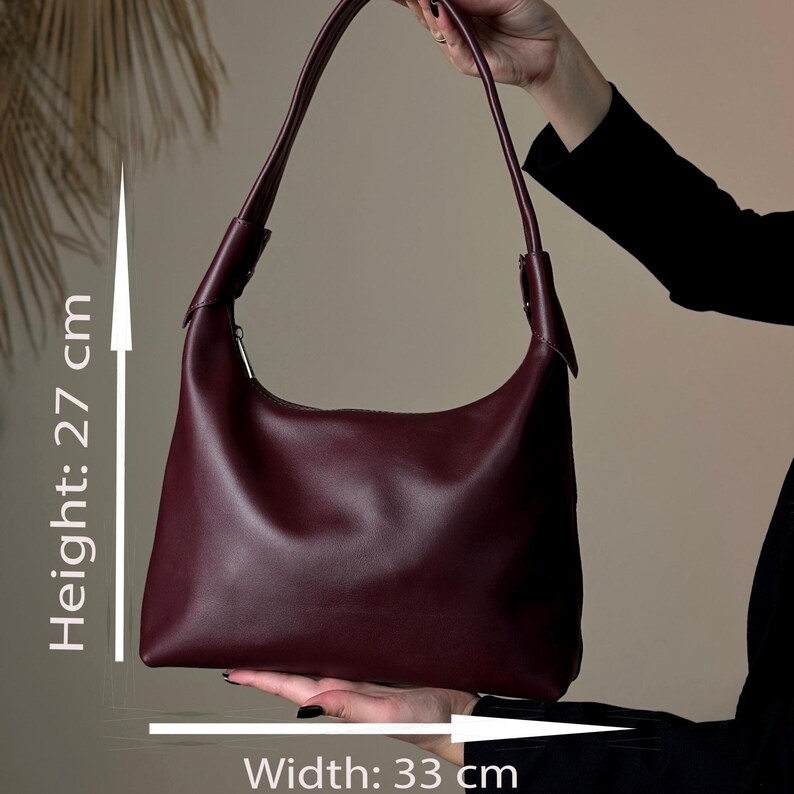 May include: A burgundy shoulder bag with a curved design. The bag is 27 cm (10.6 inches) high and 33 cm (13 inches) wide. It has a single strap and a zipper closure. The bag is held by a person.