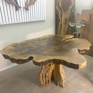 May include: A handcrafted wooden table with a natural, organic shape. The tabletop is a single slab of wood, showcasing the grain and texture. The base is made from a tree trunk, creating a rustic aesthetic. The table is approximately 122 cm long.