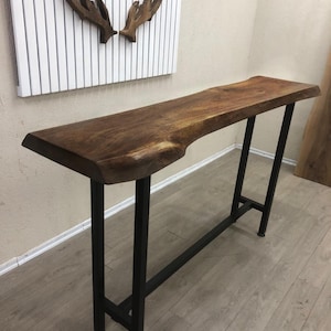 May include: A tall console table featuring a natural live edge walnut wood slab top with a rich brown grain. The table is supported by a sturdy, rectangular black metal frame base with four vertical legs and a horizontal support bar near the bottom.