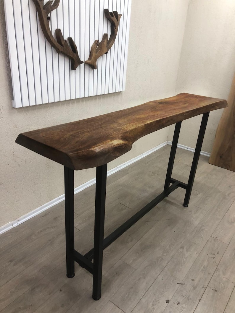 May include: A tall console table featuring a natural live edge walnut wood slab top with a rich brown grain. The table is supported by a sturdy, rectangular black metal frame base with four vertical legs and a horizontal support bar near the bottom.