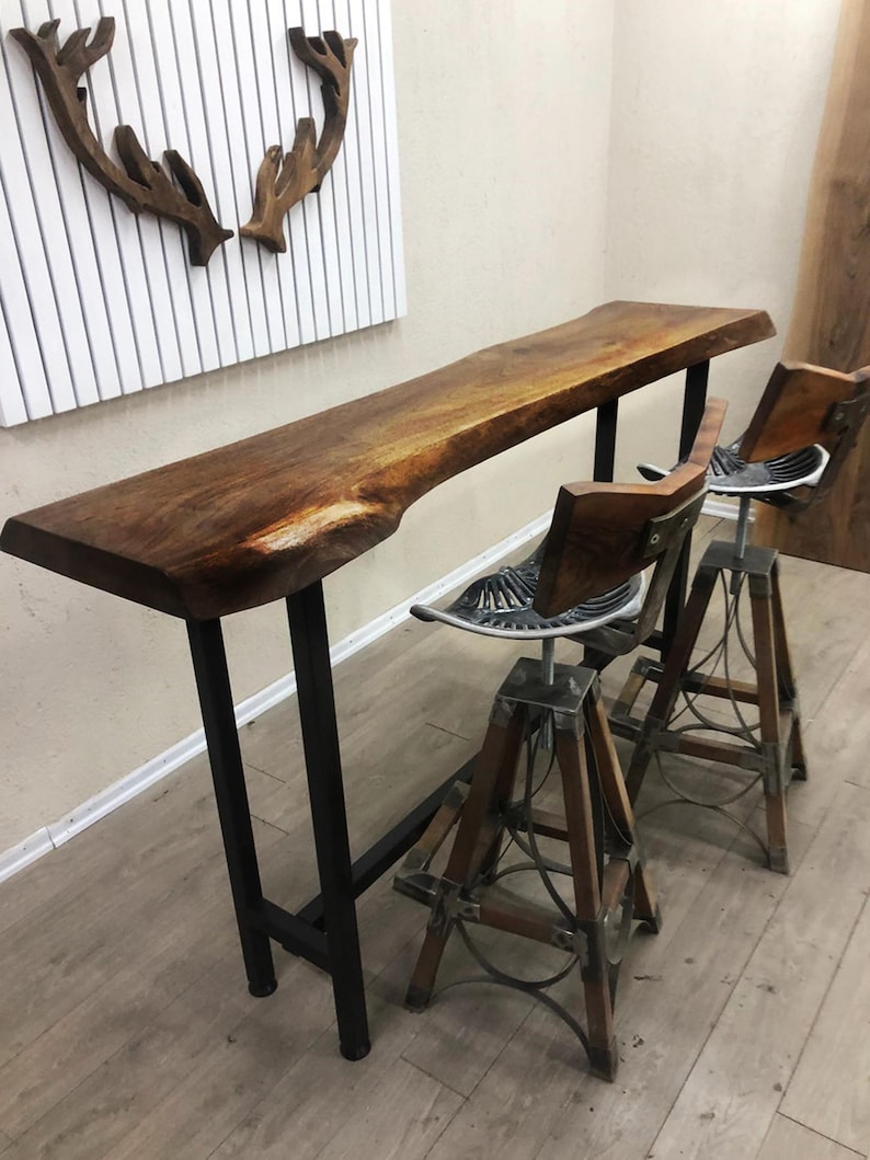 May include: A live edge wood bar table with a dark brown finish and black metal legs, accompanied by two bar stools featuring tractor-style metal seats, wooden backrests, and adjustable height bases with wooden legs and metal support rings.