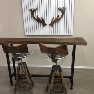 May include: A live-edge wooden bar table with a dark brown finish and black metal legs, paired with two swivel bar stools featuring wooden backrests, silver metal tractor-style seats, and tripod-style wooden and metal legs.