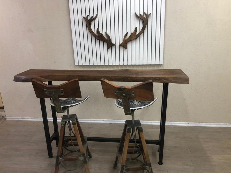 May include: A live-edge wooden bar table with a dark brown finish and black metal legs, paired with two swivel bar stools featuring wooden backrests, silver metal tractor-style seats, and tripod-style wooden and metal legs.