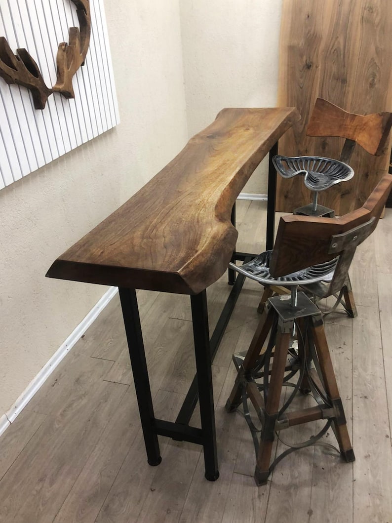 May include: A tall bar table with a natural live-edge wood top and a black metal frame, accompanied by two stools featuring tractor-style metal seats, wooden backrests, and adjustable metal bases with wooden legs and metal support accents.