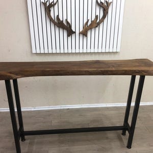 May include: A console table featuring a natural live edge dark brown wooden top with a smooth finish. The table is supported by a black metal frame consisting of four vertical legs connected by a horizontal bar near the base for added stability.
