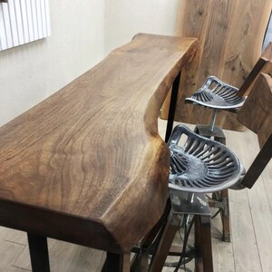 May include: A live edge wooden bar table with a natural grain finish supported by black metal legs, accompanied by two bar stools featuring silver-coloured metal tractor-style seats and wooden legs with metal cross-bracing supports.