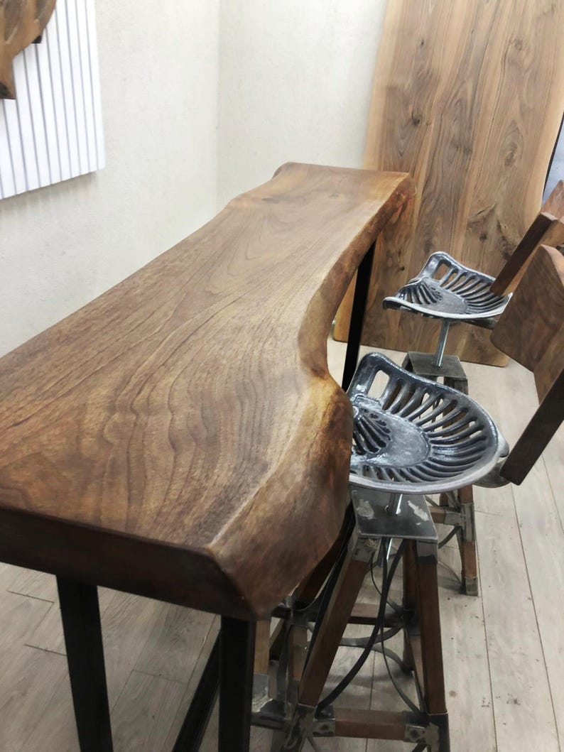 May include: A live edge wooden bar table with a natural grain finish supported by black metal legs, accompanied by two bar stools featuring silver-coloured metal tractor-style seats and wooden legs with metal cross-bracing supports.
