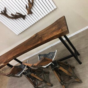 May include: A live edge wood bar table with a dark brown finish and a black metal frame, accompanied by two matching bar stools featuring wooden seats and intricate black metal bases with decorative scrollwork details and footrests.