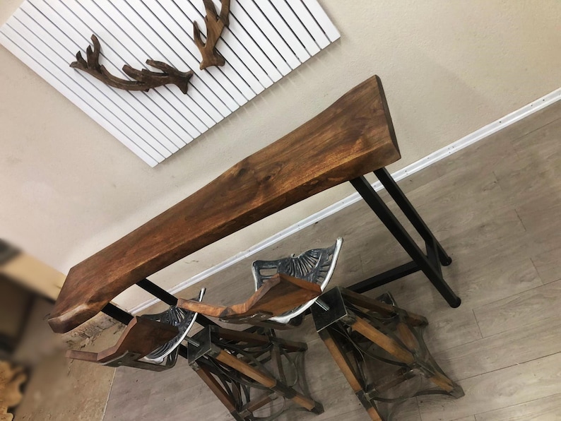 May include: A live edge wood bar table with a dark brown finish and a black metal frame, accompanied by two matching bar stools featuring wooden seats and intricate black metal bases with decorative scrollwork details and footrests.