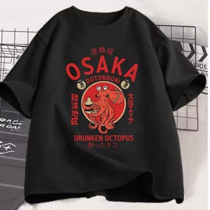 Peut inclure: T-shirt noir avec un motif de poulpe rouge et le texte "OSAKA DOTONBORI" et "DRUNKEN OCTOPUS". Le motif comprend du texte japonais et un cercle rouge autour du poulpe. Le t-shirt est de style décontracté.