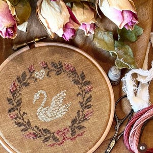 May include: A white swan cross-stitch embroidery design in a brown wooden embroidery hoop with a wreath of pink roses and green leaves. The hoop is surrounded by dried roses, a thimble, scissors, and embroidery floss.