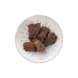May include: A white ceramic plate with a gold rim and gold starburst design holds several pieces of dark brown, textured food. The food pieces vary in size and shape, with a rough, uneven surface.