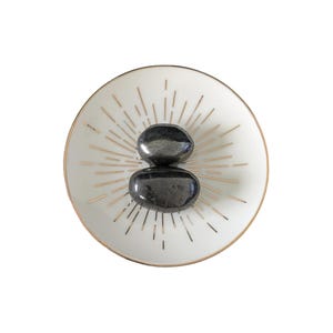 May include: A white ceramic dish with a gold rim and gold radiating lines. Two dark grey, oval-shaped stones are stacked on top. The dish has a minimalist design, suitable for decorative purposes or as a small trinket tray.