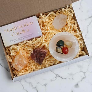 May include: A gift box containing crystals, a small bowl with stones, and a card that reads "Manifestation Candles". The box is filled with yellow paper filler. The crystals are various colours, including orange, pink, and red.