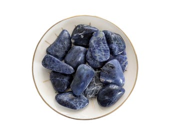 May include: A collection of polished, tumbled sodalite stones in shades of deep blue, navy, and white, arranged in a circular white ceramic dish with a thin gold-colored rim. The stones have smooth, glossy surfaces with visible mineral veins.