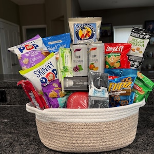Könnte beinhalten: Ein geflochtener Seilkorb mit weißem Oberteil und beigem Unterteil, gefüllt mit verschiedenen Snacks wie SkinnyPop-Popcorn, Pirate's Booty, Cheez-It-Crackern, Flipz-Brezeln, Annie's-Snacks, zwei Celsius-Energydrinks, einer Lepro-Taschenlampe und 5-Kaugummi.