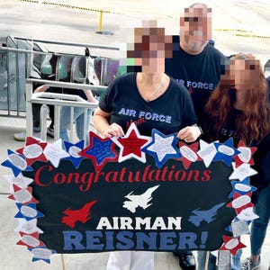 May include: A black sign with the words "Congratulations" and "Airman Reisner!" in red and blue. The sign is decorated with red, white, and blue star-shaped cutouts. The sign is held by a person wearing an "Air Force Mom" t-shirt.