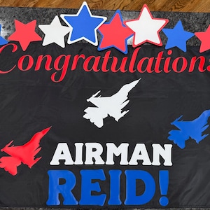 May include: A black rectangular banner featuring the red text Congratulations, the white text AIRMAN, and the blue text REID! with an exclamation point. The banner is decorated with red, white, and blue star cutouts and three fighter jet silhouettes.