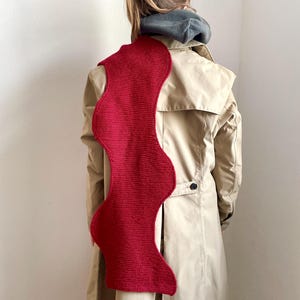 May include: A red knitted scarf with a wavy design. The scarf is worn by a person wearing a beige trench coat.