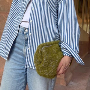 Könnte beinhalten: Eine flauschige, olivgrüne Strick-Clutch, gehalten vor einem weißen T-Shirt, einem blau-weiß vertikal gestreiften Hemd und einer hellblauen Jeans. Die Tasche zeichnet sich durch eine weiche, strukturierte Garnbeschaffenheit und einen einfachen Verschluss aus.