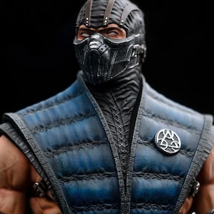 May include: A detailed action figure of a character from the Mortal Kombat video game series. The figure has a black mask and helmet, a blue and black quilted vest, and a silver emblem. The figure is set against a black background.