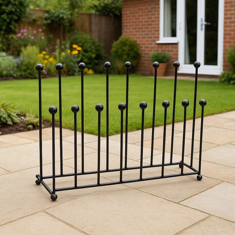 May include: Black metal boot rack with a series of vertical rods topped with round spheres. The rack is designed to hold multiple pairs of boots. The rack is placed on a stone patio with a green lawn and garden in the background.