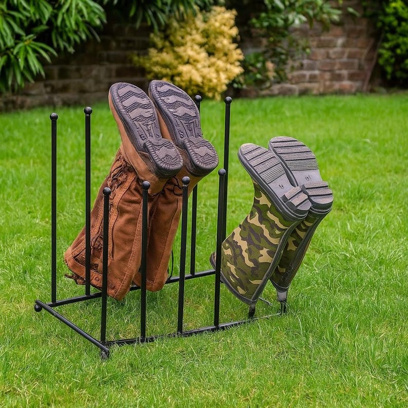 May include: A black metal boot rack holding two pairs of boots. One pair is brown leather, and the other is camouflage. The rack is on a green lawn, with a brick wall and bushes in the background.